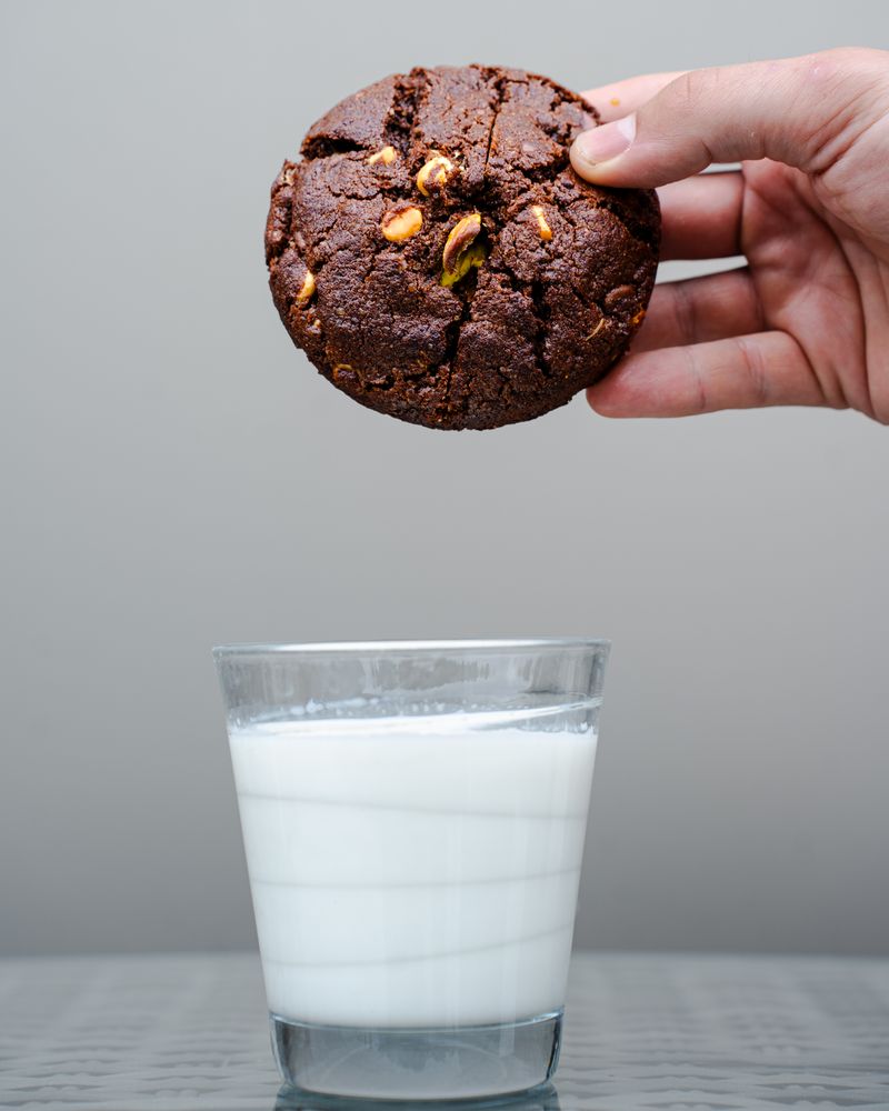 Hand holding a Dubai-style cookie with pistachio nuts above a glass of milk - ready to dunk
