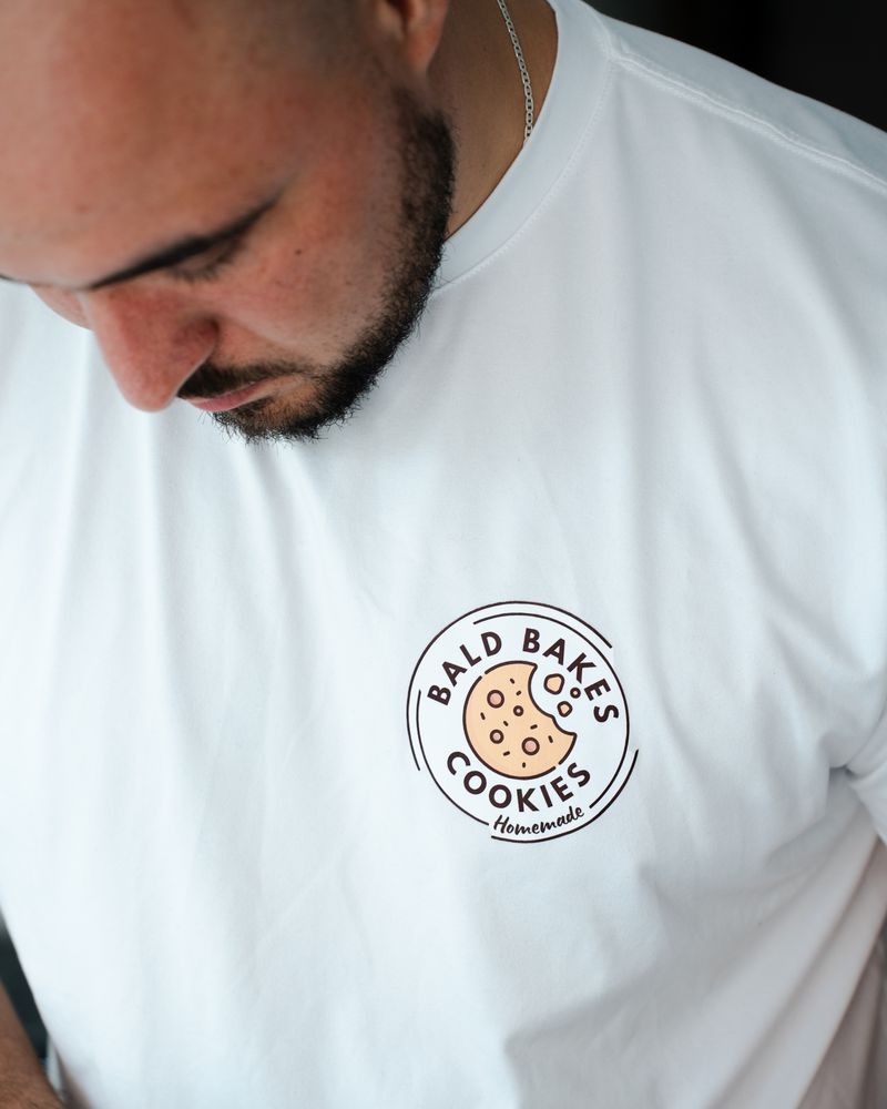 The bald baker wearing a white t-shirt with 'Bald Bakes Cookies' logo